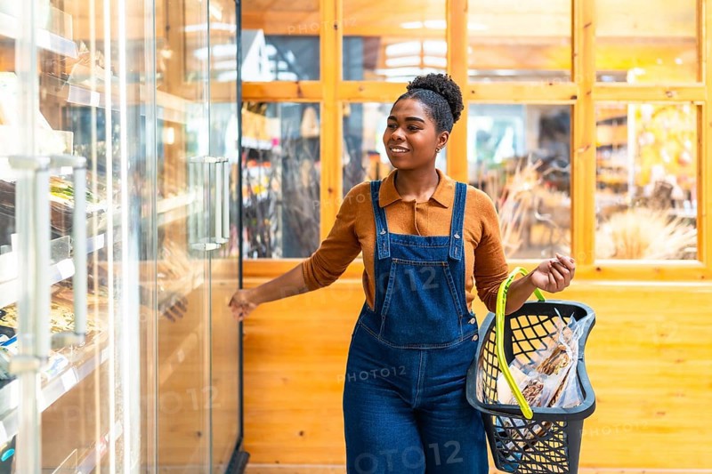 Smiling black woman opening refrigerated section door in grocery store holding shopping basket