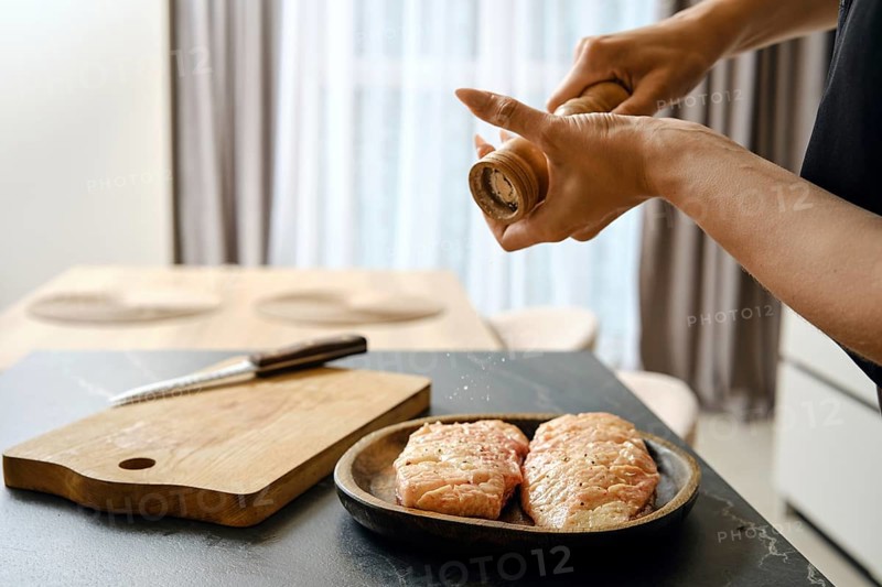 A person seasons fresh duck fillets with sea salt in a stylish kitchen. The wooden cutting board and knife are nearby, while natural light brightens the space from the window