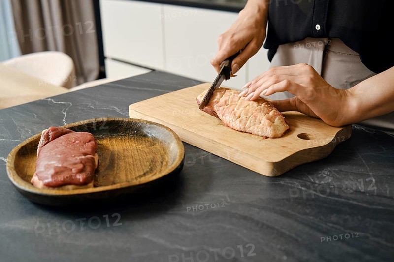 A person skillfully cuts the skin crosswise on a duck breast on a wooden board placed on a dark countertop. Another piece of meat rests on a wooden plate nearby