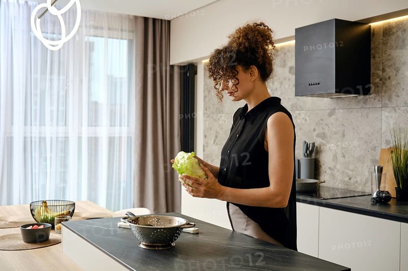 In a contemporary kitchen filled with natural light, a woman holds a head of lettuce while preparing to create a fresh salad. Various fruits and vegetables are seen nearby
