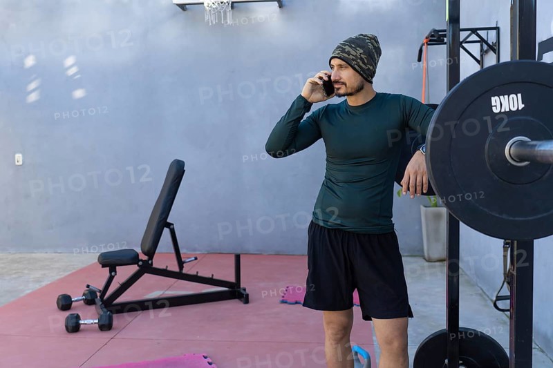 Athlete taking a break from an intense workout, leaning against a weightlifting rack while engaged in a conversation on his mobile phone in a home gym setting