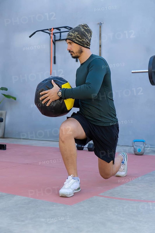 Athlete wearing a beanie, long-sleeved shirt, and shorts performs lunges while holding a medicine ball in an outdoor gym setting, demonstrating strength and balance