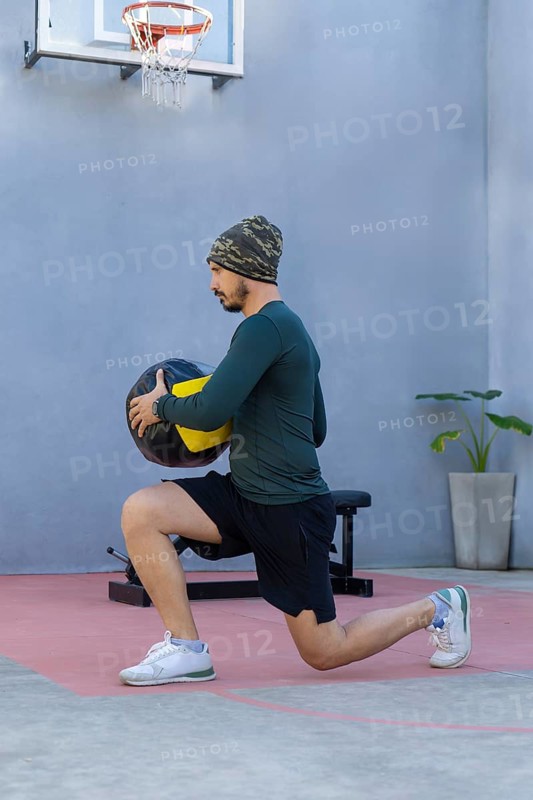 Young sportsman wearing a beanie and sportswear is lunging with a medicine ball in an outdoor gym, positioned near a basketball hoop in an urban setting