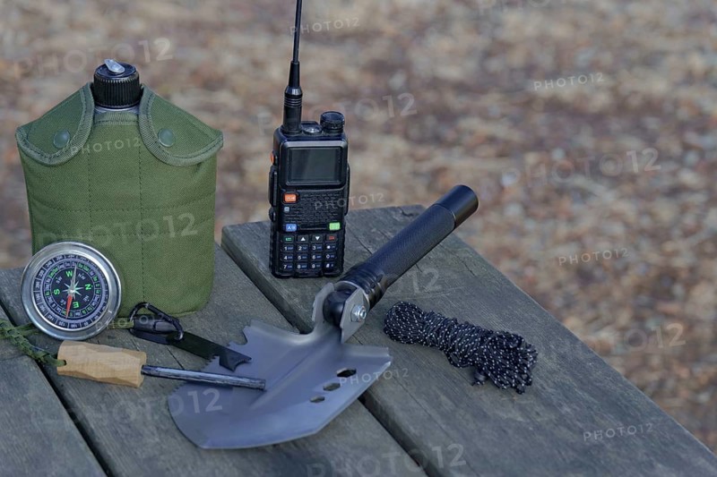 Essential survival gear including walkie talkie, compass, folding shovel, and fire starter are arranged on a wooden table in nature