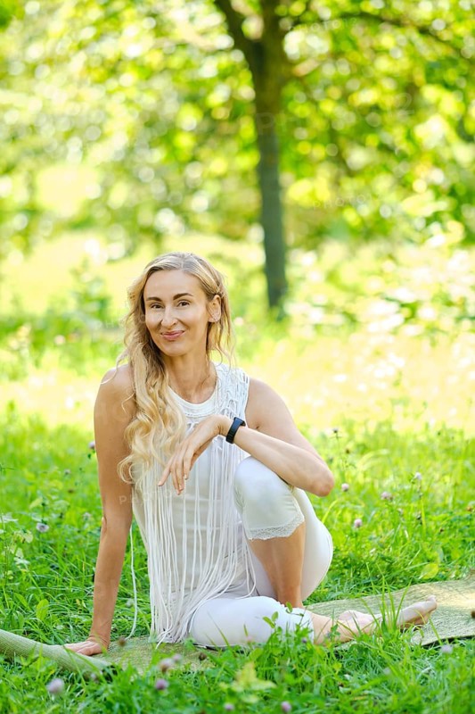A woman dressed in white sits gracefully on a mat in a lush park, surrounded by vibrant green grass and blooming flowers. She enjoys a peaceful moment during her yoga practice under the warm sun