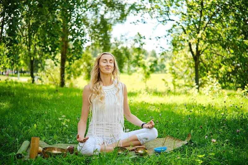 A woman sits cross-legged on a mat in a lush green park, eyes closed and arms resting on her knees. She focuses on mindfulness and tranquility, surrounded by trees and nature