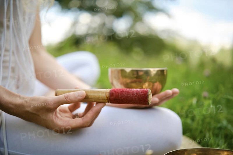 A person is holding a red-tipped mallet next to a singing bowl, seated on grass in a serene outdoor setting. This meditative activity takes place in a tranquil environment, encouraging relaxation