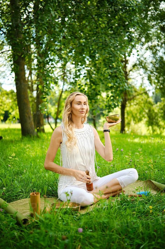 A woman practices meditation outdoors, holding a singing bowl while seated on a mat in a field. The surrounding nature and sunlight enhance the calming atmosphere of the moment