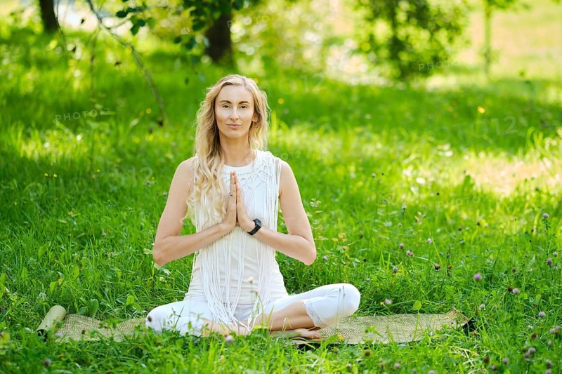 A woman dressed in white sits cross-legged on a mat, practicing yoga outdoors. Surrounded by vibrant green grass and trees, she has her hands in a prayer position, embodying peace and tranquility