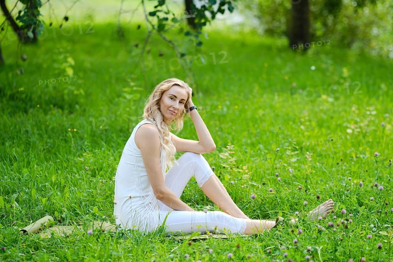 A young woman sits comfortably on a blanket in a lush green park. She has wavy hair and wears a white outfit