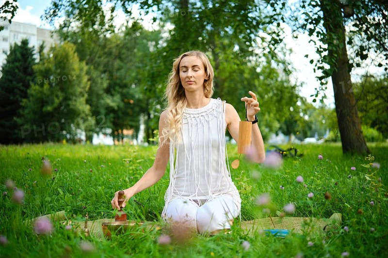 A person sits peacefully on a mat in a green park, gently playing singing bowls during meditation. The sun filters through the trees, creating a serene atmosphere as nature surrounds them