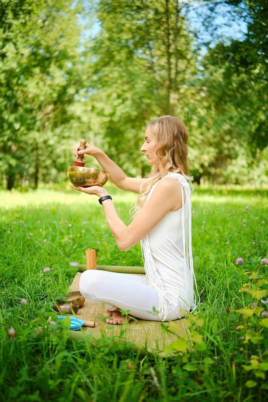 A woman sits cross-legged on a mat in a lush green field, playing a singing bowl with a mallet. Sunlight filters through the trees as she engages in meditation and mindfulness