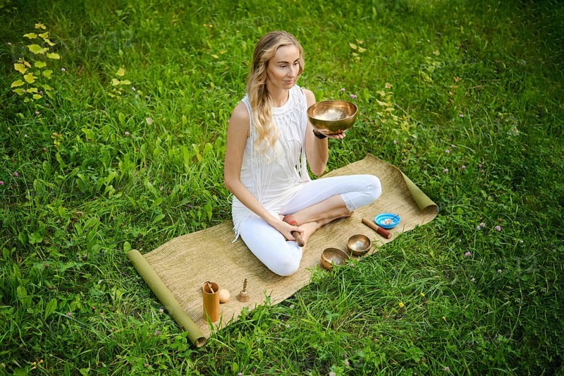 A woman sits cross-legged on a mat in a lush green field, holding a singing bowl. She is fully engaged in a meditative practice, surrounded by additional bowls and calming symbols