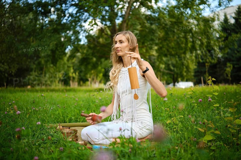 A woman sits cross-legged on the grass, surrounded by greenery, gently striking singing bowls with mallets