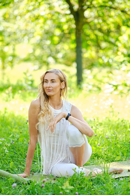 A woman with long hair sits elegantly in a vibrant green park. Dressed in white, she poses thoughtfully amidst flowers and sunshine while enjoying the peaceful atmosphere