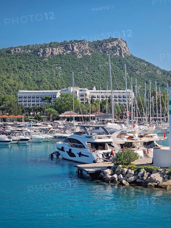 Kemer marina filled with numerous boats and yachts with Calis Hill in the background. A lighthouse stands prominently on the rocky pier, overlooking the clear blue water under a bright blue sky
