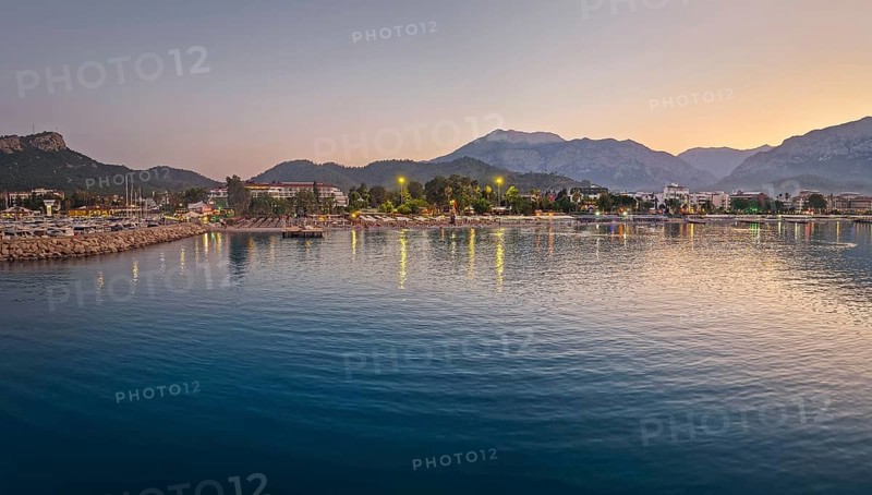 Dusk scene in Kemer, Turkey, showing the town illuminated coastline and beach, reflected beautifully on the calm sea. Majestic Taurus mountains silhouettes in the background