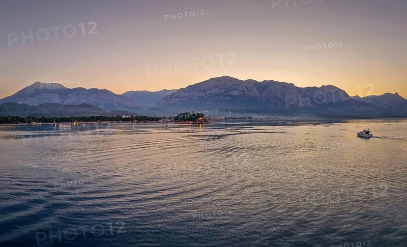 Seascape sunset panorama over the Mediterranean Sea near Kemer, Turkey. Panoramic view to the moonlight beach at dusk and a lone boat cruises across the calm water towards the majestic mountains