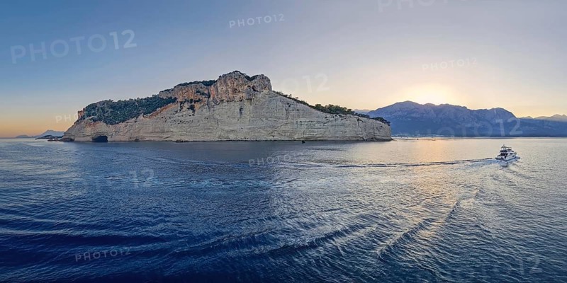 Vibrant evening seascape with a boat cruising away on the blue sea, near the Pirate Cave in Kemer at sunset. Beautiful Turkish mediterranean coastline lined with tree covered rocky cliffs at dusk