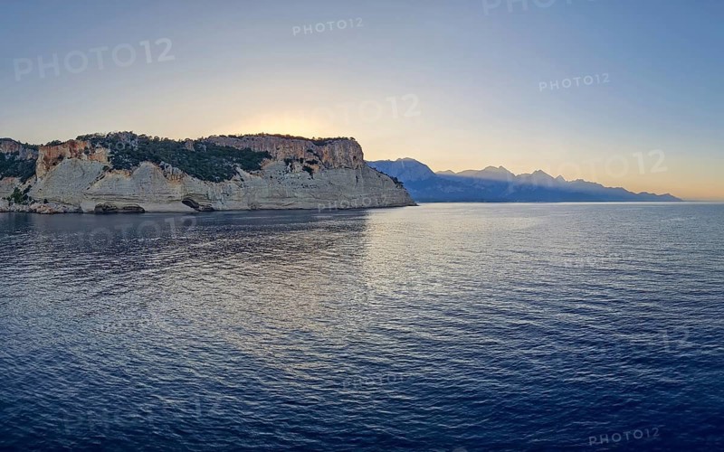 Vibrant seascape sunset with a view to the Pirate Cave in Kemer, Turkey. Beautiful Turkish mediterranean coastline lined with tree covered rocky cliffs and hills under a clear evening sky