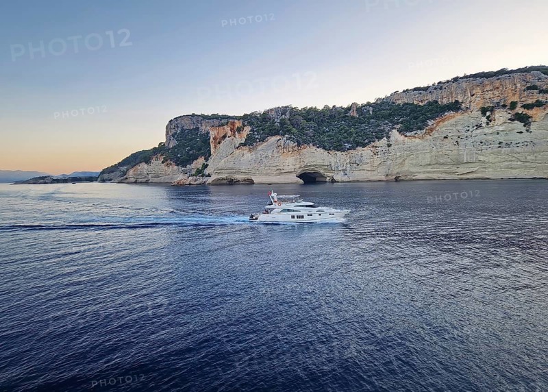 Vibrant evening seascape with a boat cruising away on the blue sea, near the Pirate Cave in Kemer at sunset. Beautiful Turkish mediterranean coastline lined with tree covered rocky cliffs at dusk