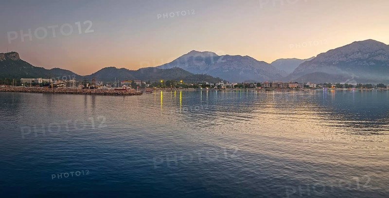 Dusk scene in Kemer, Turkey, showing the town illuminated coastline and beach, reflected beautifully on the calm sea. Majestic Taurus mountains silhouettes in the background