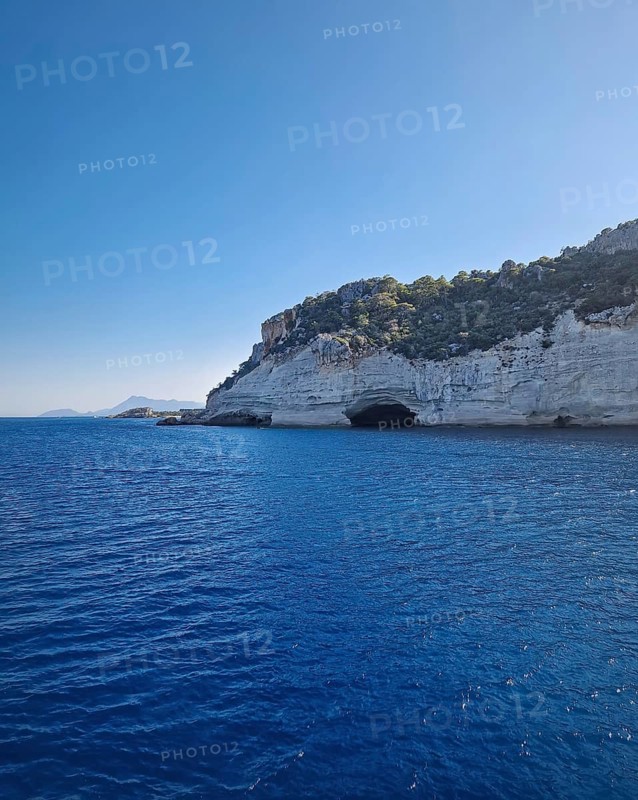 Vibrant seascape with the Pirate Cave in Kemer, Turkey. Beautiful Turkish mediterranean coastline lined with tree covered rocky cliffs and hills under a clear sky
