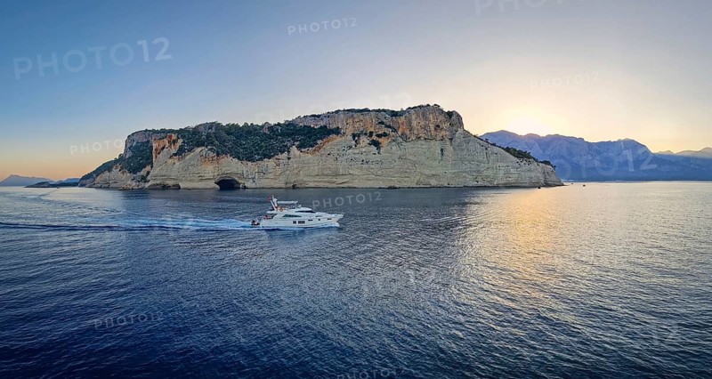 Evening seascape panorama with a boat cruising away on the blue sea, near the Pirate Cave in Kemer at sunset. Beautiful Turkish mediterranean coastline lined with tree covered rocky cliffs at dusk