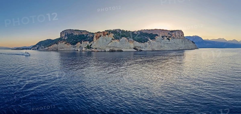 Evening seascape panorama with a boat cruising away on the blue sea, near the Pirate Cave in Kemer at sunset. Beautiful Turkish mediterranean coastline lined with tree covered rocky cliffs at dusk