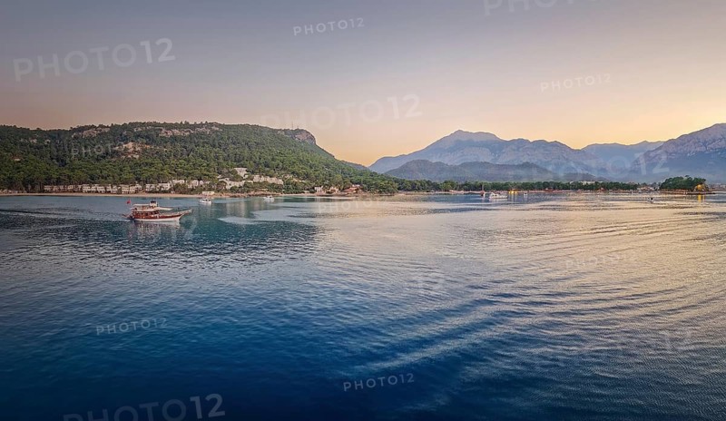 Seascape sunset panorama over the Mediterranean Sea near Kemer, Turkey. Panoramic view to the moonlight beach at dusk and a lone boat cruises across the calm water towards the majestic mountains