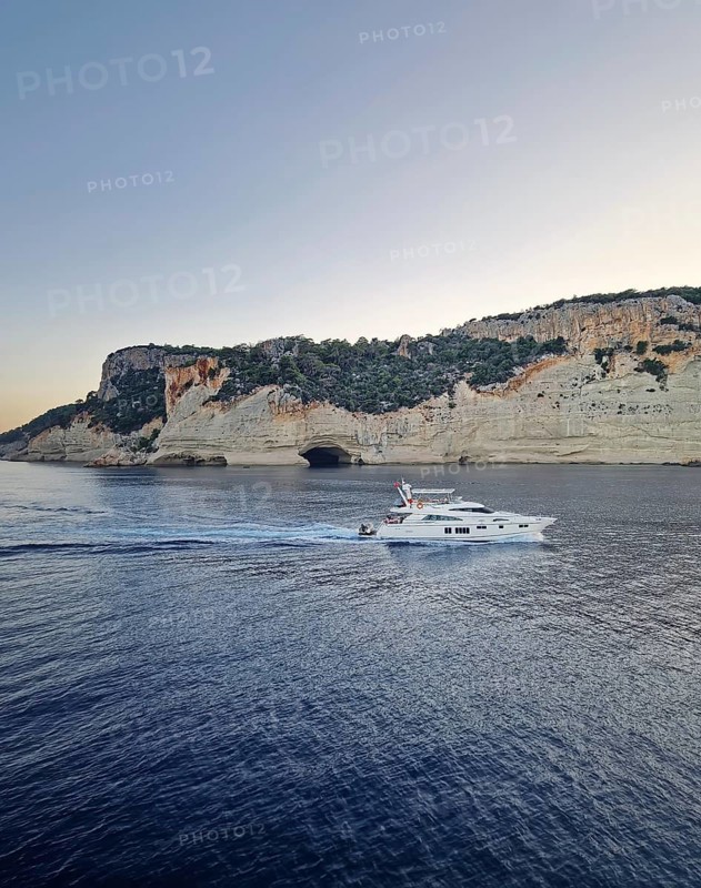 Vibrant evening seascape with a boat cruising away on the blue sea, near the Pirate Cave in Kemer at sunset. Beautiful Turkish mediterranean coastline lined with tree covered rocky cliffs at dusk