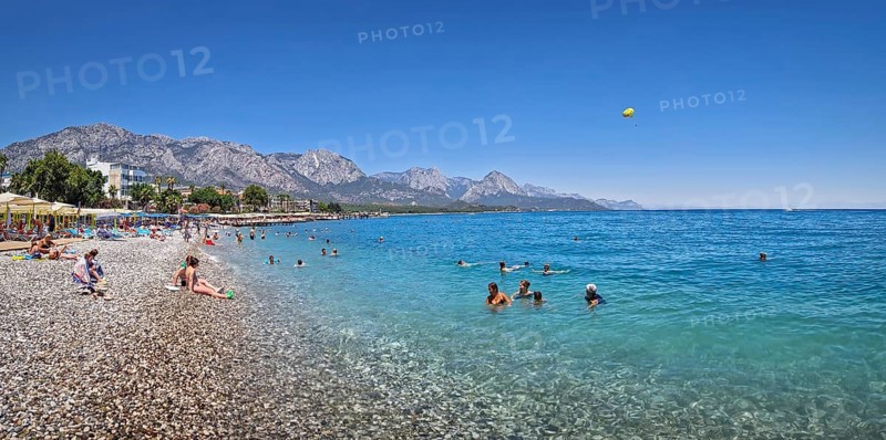 KEMER, TURKEY - JULY 5, 2025 Kemer municipality public beach with vibrant pebble under a clear, turquoise sea water. People swimming and sunbathing on the shore. Beautiful Taurus mountains at horizon