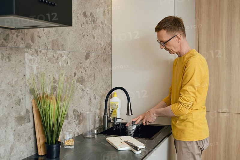 A person in a yellow long-sleeve shirt is washing dishes at a sleek kitchen sink. Sunlight illuminates the modern kitchen with stone and wood elements, creating a warm atmosphere