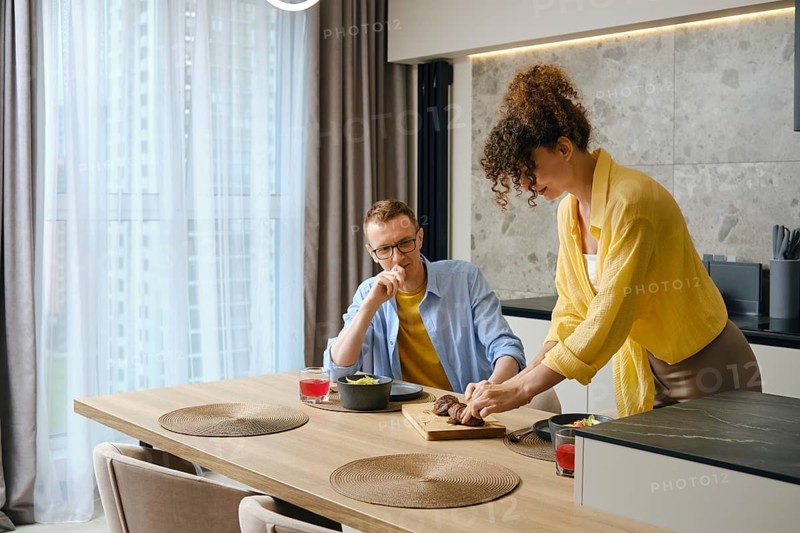 A man and woman are preparing a meal together in a modern kitchen. The woman slices food on a wooden cutting board while the man observes, smiling