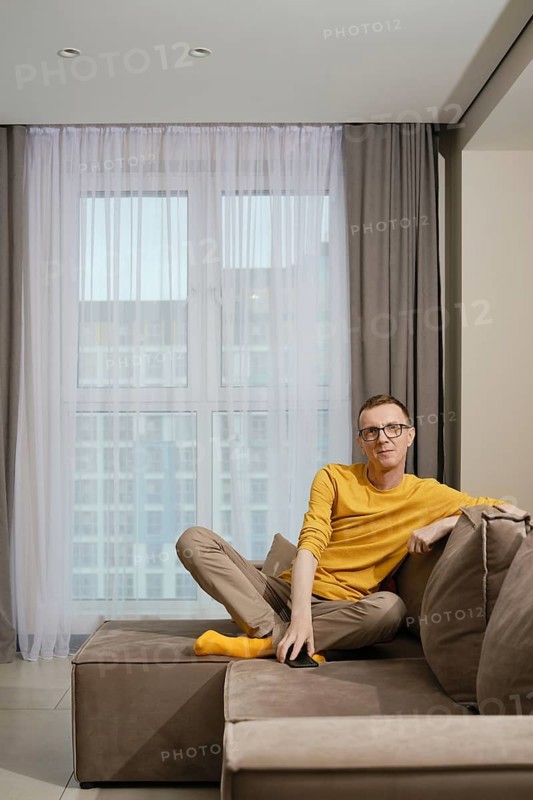 A man wearing a yellow shirt and socks and beige pants sits comfortably on a brown couch, relaxed in a well-lit modern apartment with large windows and curtains
