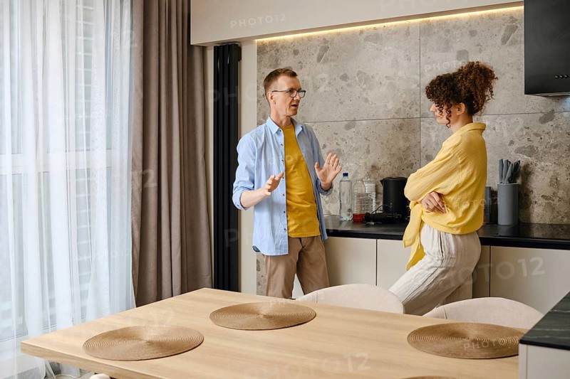 A man and a woman are discussing animatedly in a stylish kitchen, with sunlight streaming through the window. The atmosphere feels relaxed as they share ideas, leaning against the countertops