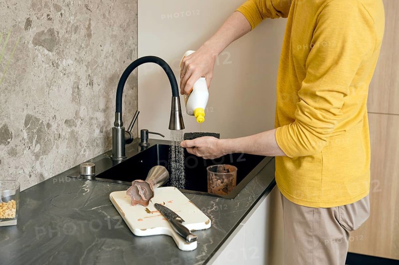 A person wearing a yellow shirt is pouring liquid soap on sponge to clean kitchen utensils and a cutting board in a dark kitchen sink