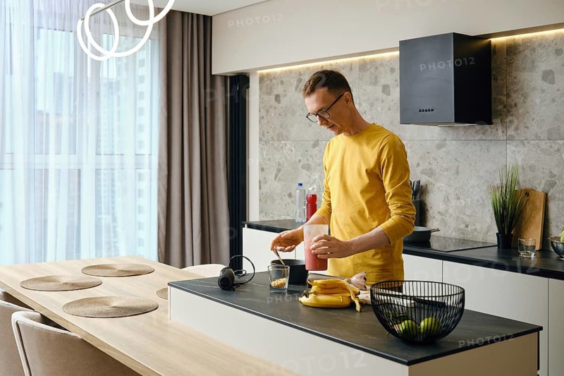In a sleek kitchen, a man in a yellow shirt blends ingredients for a smoothie. He stands at a well-lit counter with fruits, a glass, and kitchen appliances around him, engaged in healthy cooking