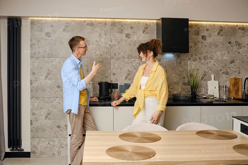 A man and a woman share an animated discussion in a contemporary kitchen during the day. The kitchen features sleek design elements and natural light highlighting their interaction