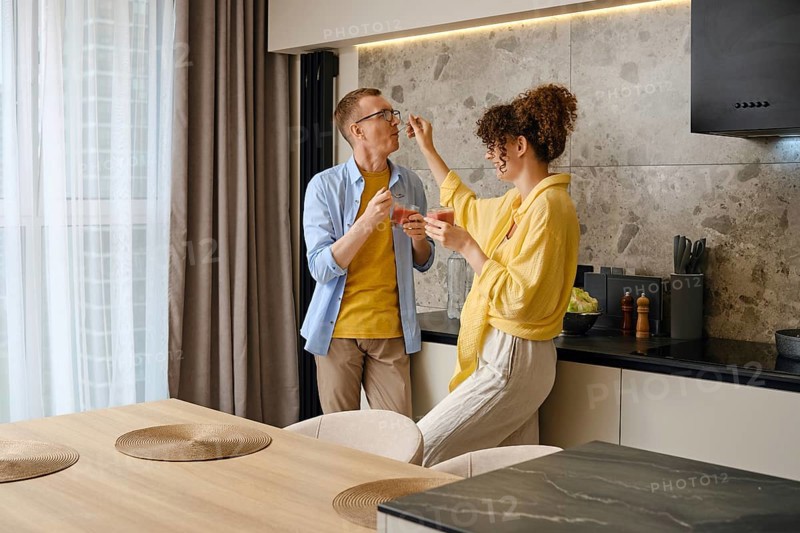 A couple shares a joyful moment in their stylish kitchen. Wife playfully feeds her husband with a spoon