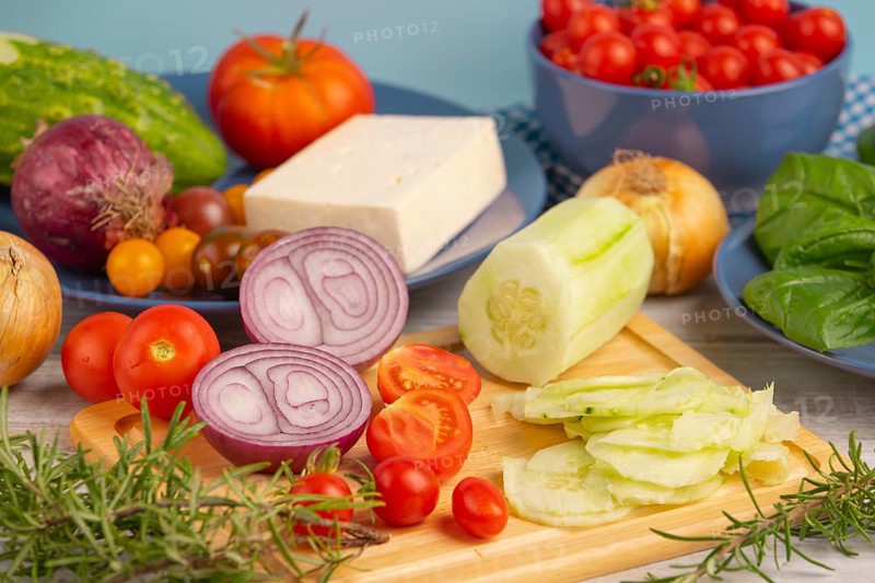 Fresh and sliced vegetables with feta on a wooden board, surrounded by basil and rosemary, rustic cuisine