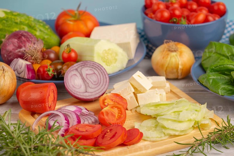 Fresh and sliced vegetables with feta on a wooden board, surrounded by basil and rosemary, rustic cuisine