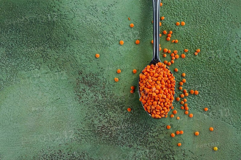 Bright orange lentils spill from a silver spoon onto a textured green surface, highlighting the vibrant colors and nutritional value of this versatile ingredient in cooking