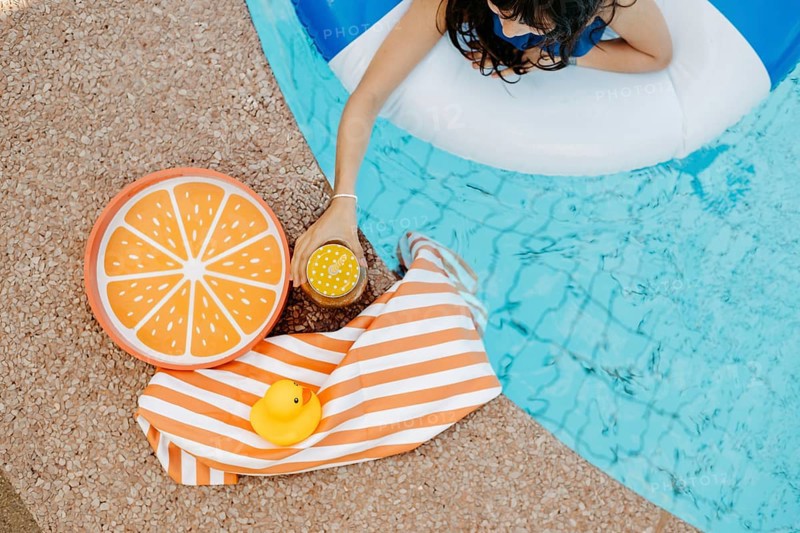 Girl with a swimming tyre in the pool, a striped bath towel, a swimming duck, a cocktail and an orange tray at the edge of the pool