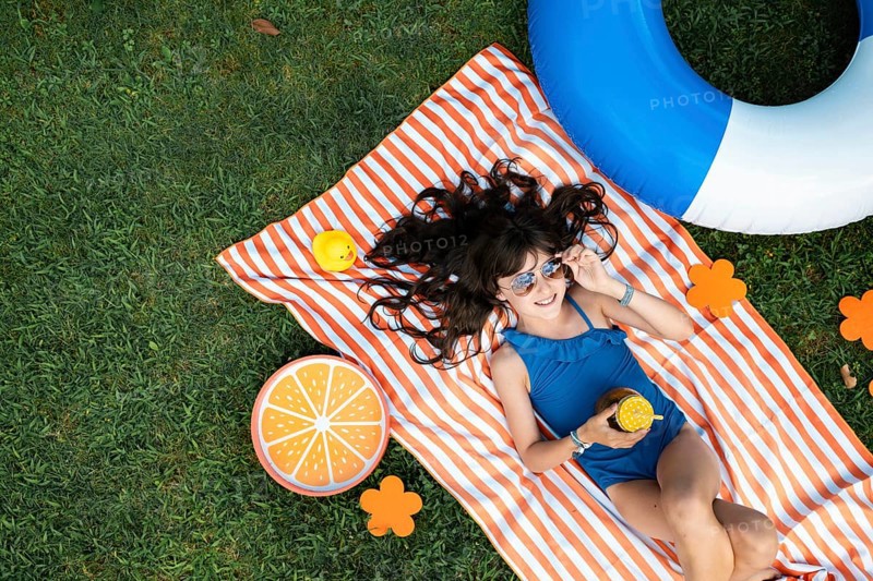 Girl, 10, with sunglasses in a blue swimming costume, lying relaxed in the meadow with a cocktail on a towel
