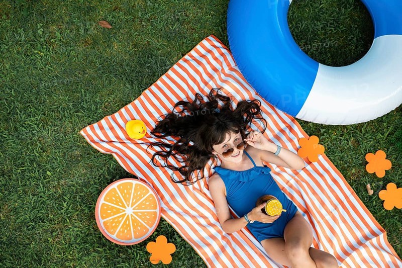 Girl, 10, with sunglasses in a blue swimming costume, lying relaxed in the meadow with a cocktail on a towel