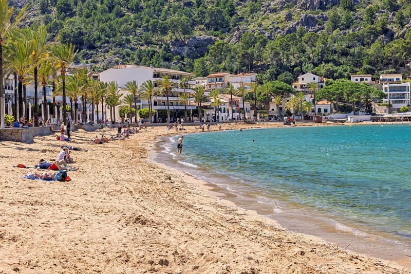 Sand beach with tourists at a tourist resort with houses at the shore line and palm trees a sunny summer day, Port de Sóller, Mallorca, Spain