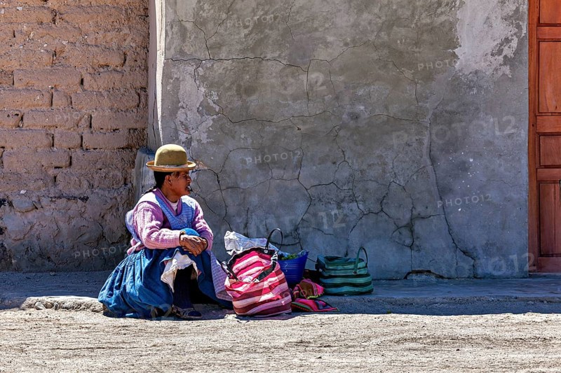Woman in traditional dress sitting on the floor next to colourful bags in front of a wall, Traditional woman in a village in the Salar de Uyuni in Bolivia