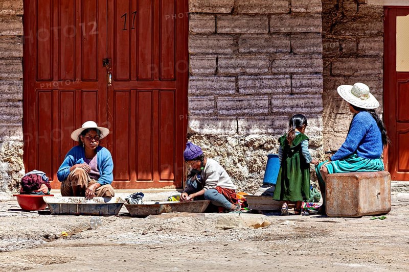 Women and children sit in front of a stone house, busy with housework. The sun shines brightly, traditionally dressed woman and children sit on the ground in the Salar de Uyuni in Bolivia