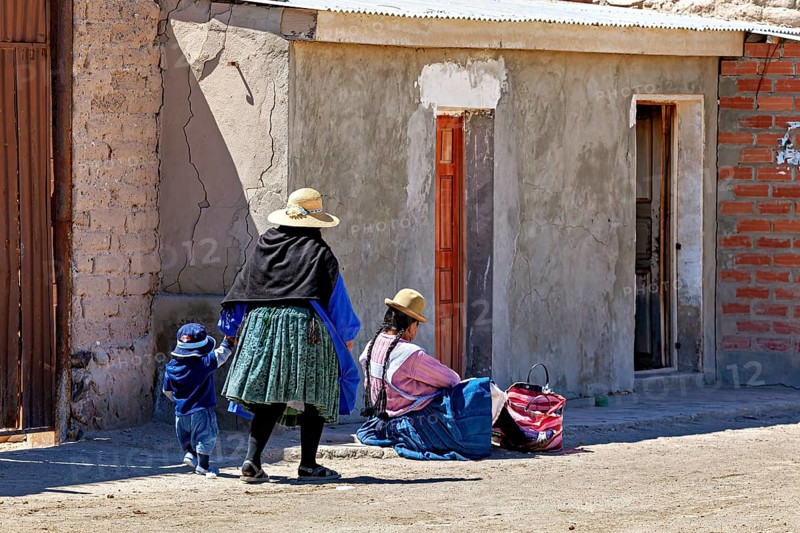 Woman and child in traditional dress standing in front of a brick house on a sunlit street, Traditional woman in a village in the Salar de Uyuni in Bolivia